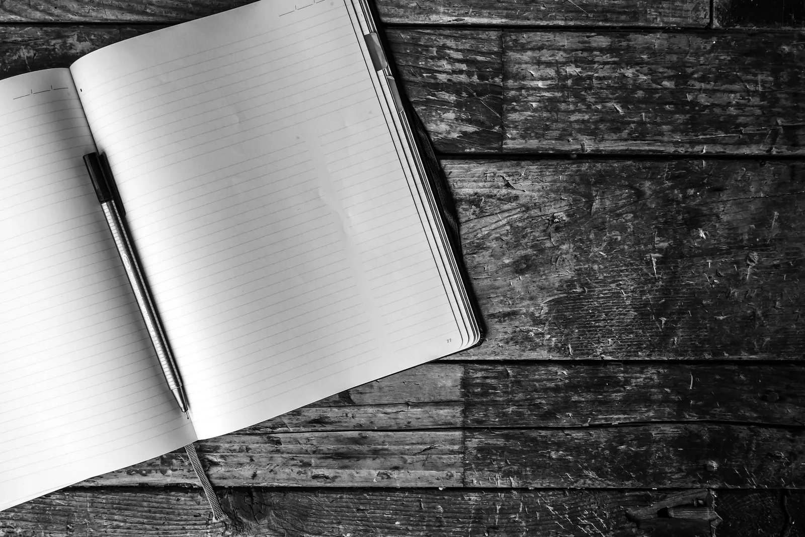 An open notebook and a pen resting on a wooden desk, in black and white.