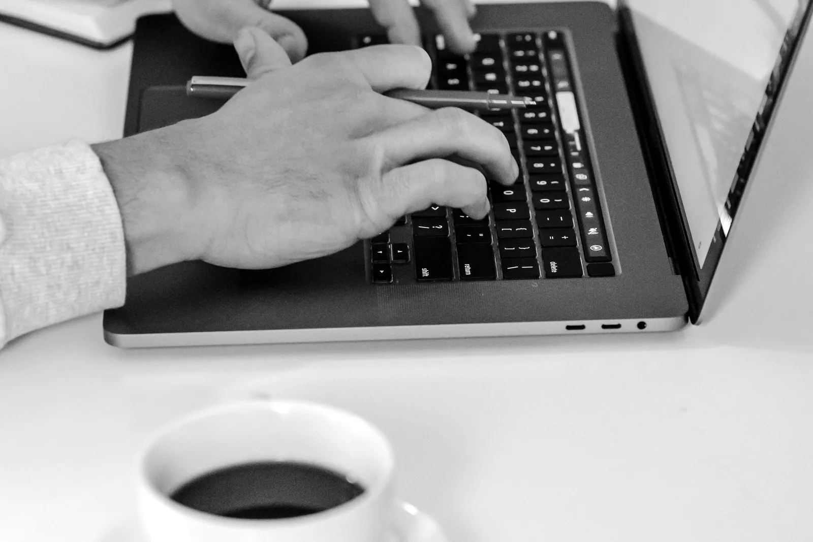 Hands typing on a laptop beside a cup of coffee on a café table, in black and white.