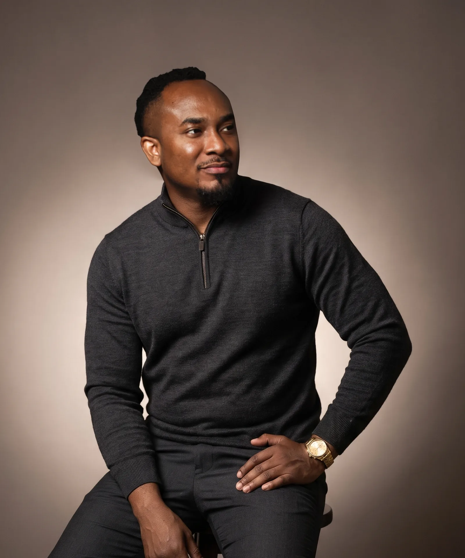 Kemal Brown in a charcoal quarter-zip, seated on a stool, smiling and looking aside.