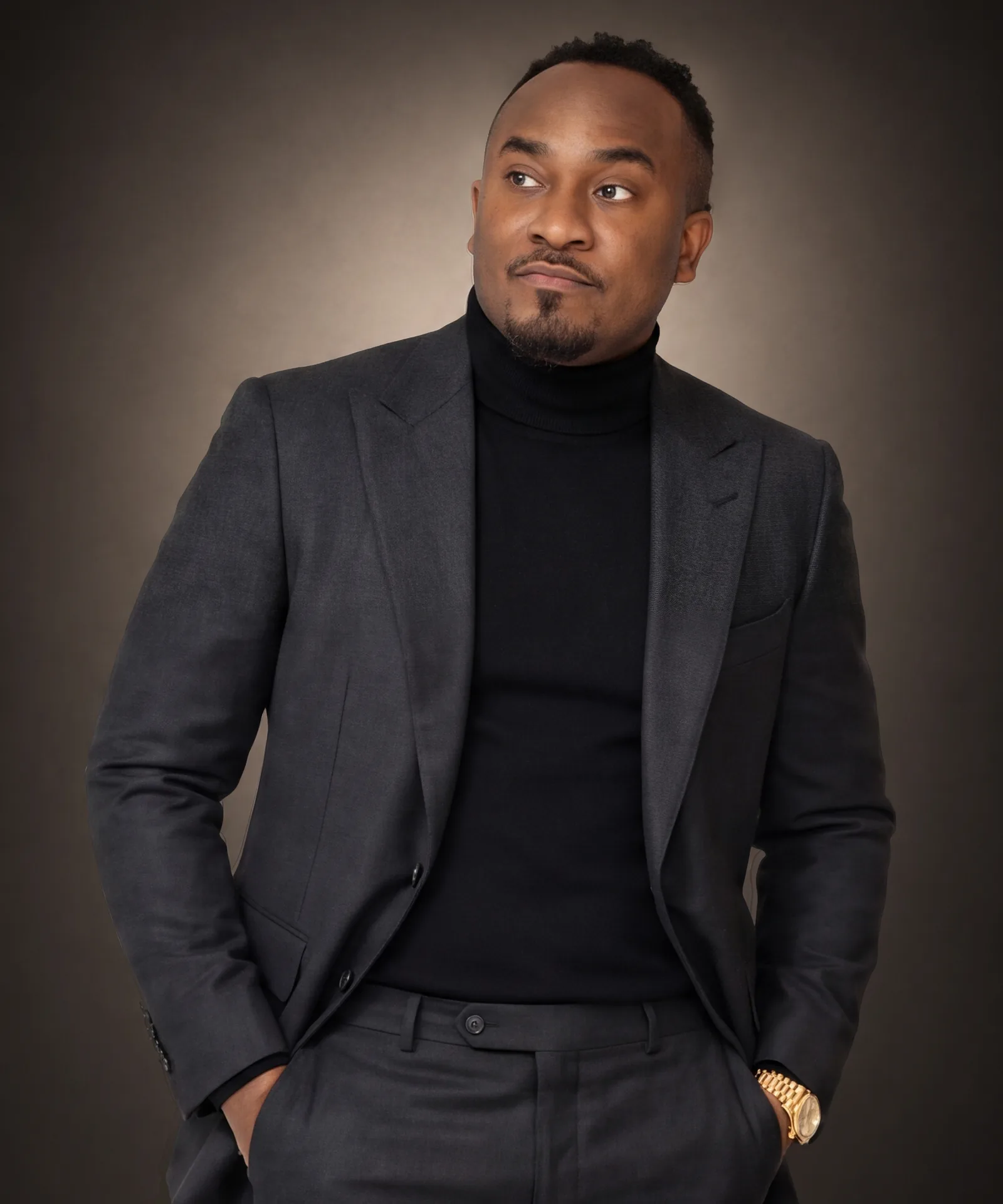 Kemal Brown in a charcoal suit and black turtleneck, hands in his pockets, looking to the side.