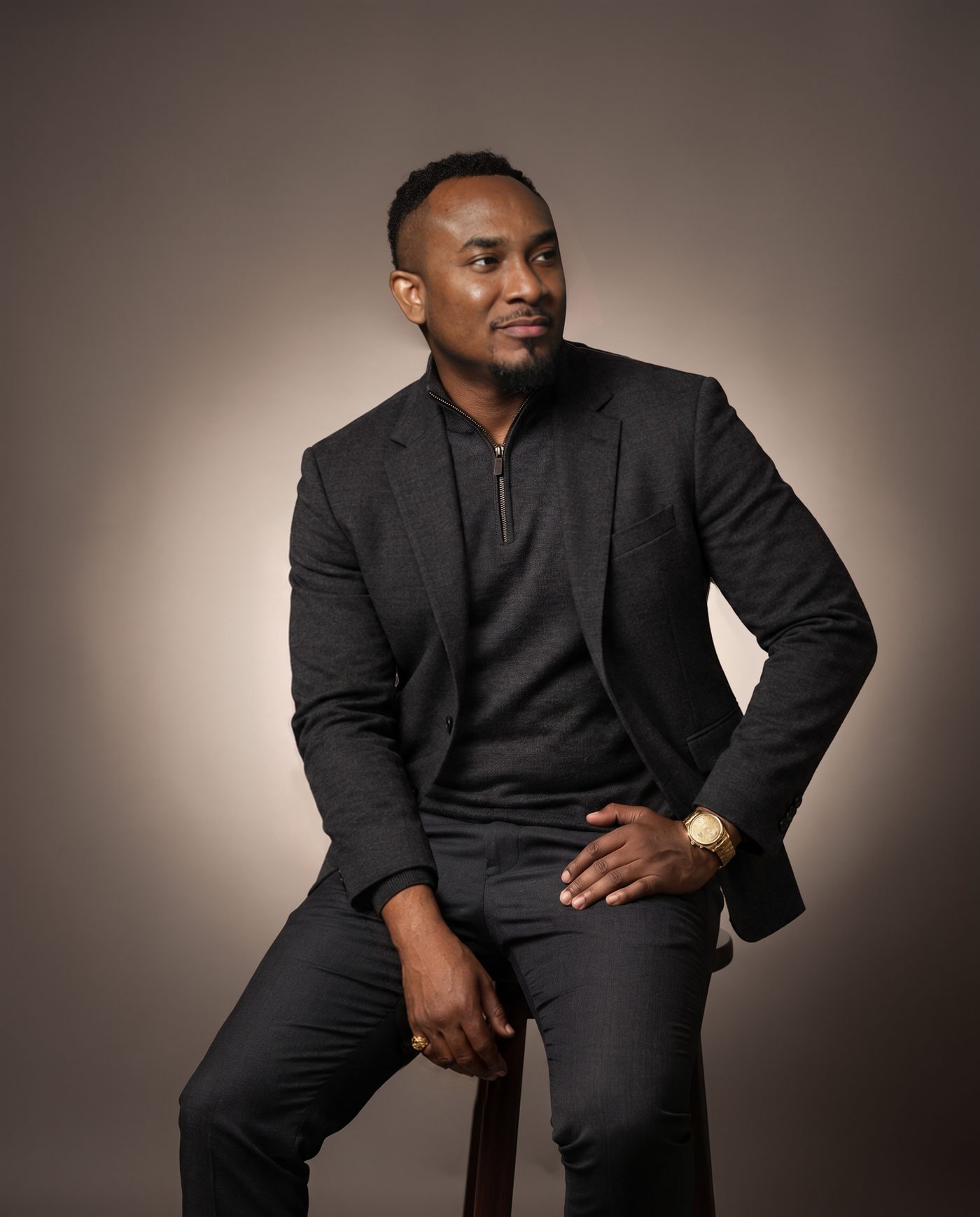 Kemal Brown in a charcoal blazer over a quarter-zip, seated on a stool, smiling and looking aside, gold watch on his wrist.