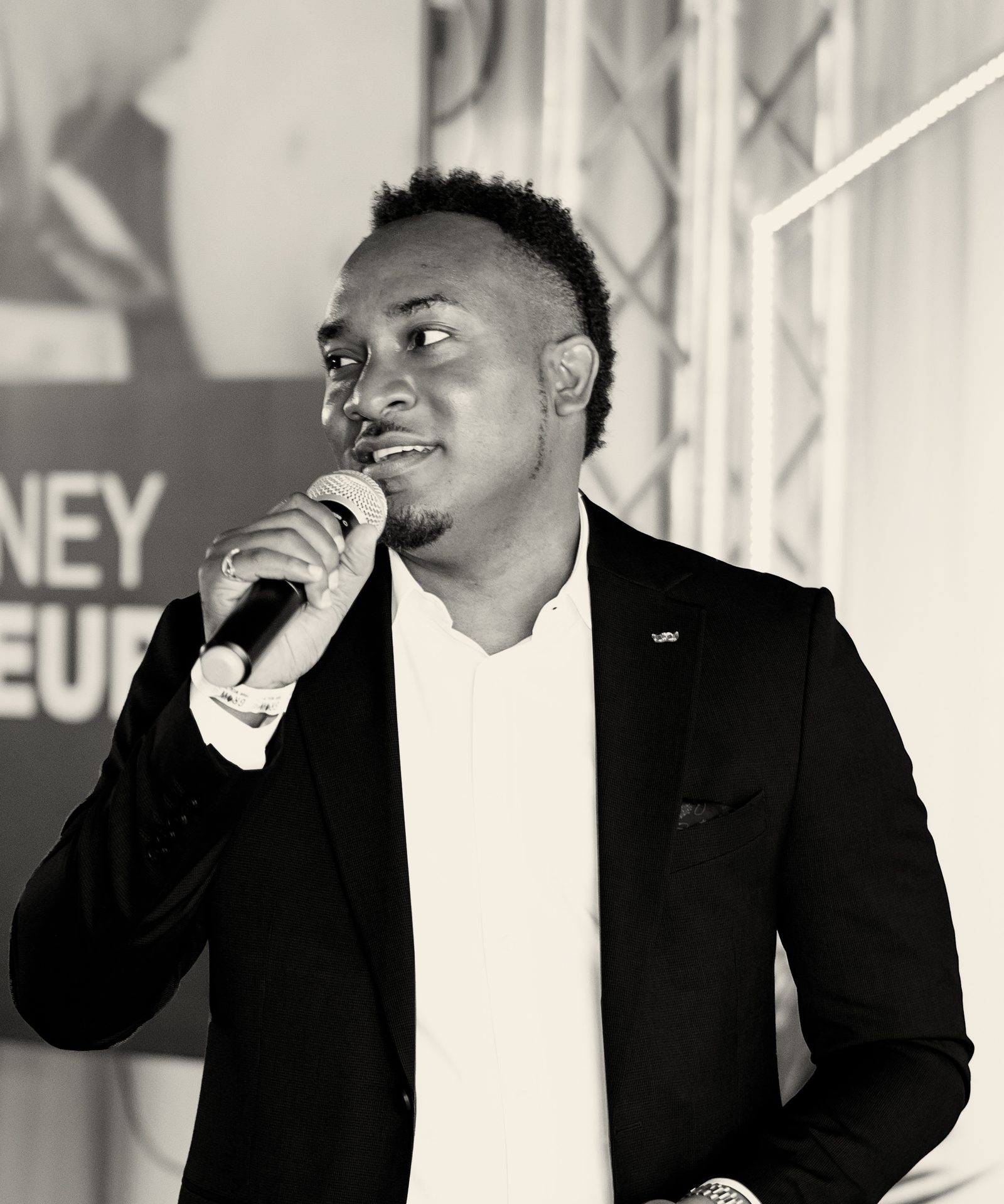 Kemal Brown standing on stage, speaking into a handheld microphone, in a black blazer and white shirt.