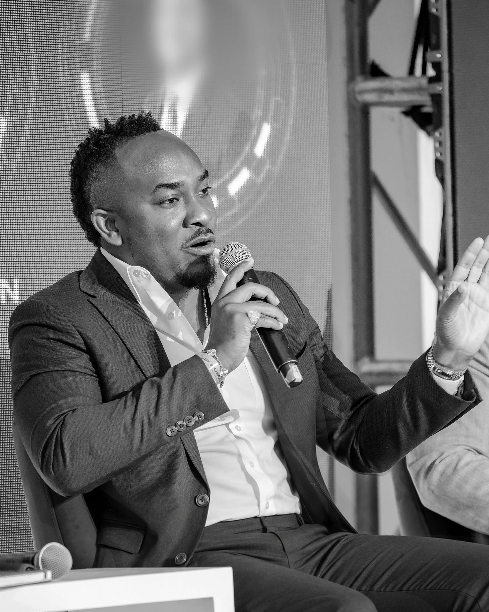 Kemal Brown seated on a conference panel, holding a microphone and gesturing as he speaks, in black and white.