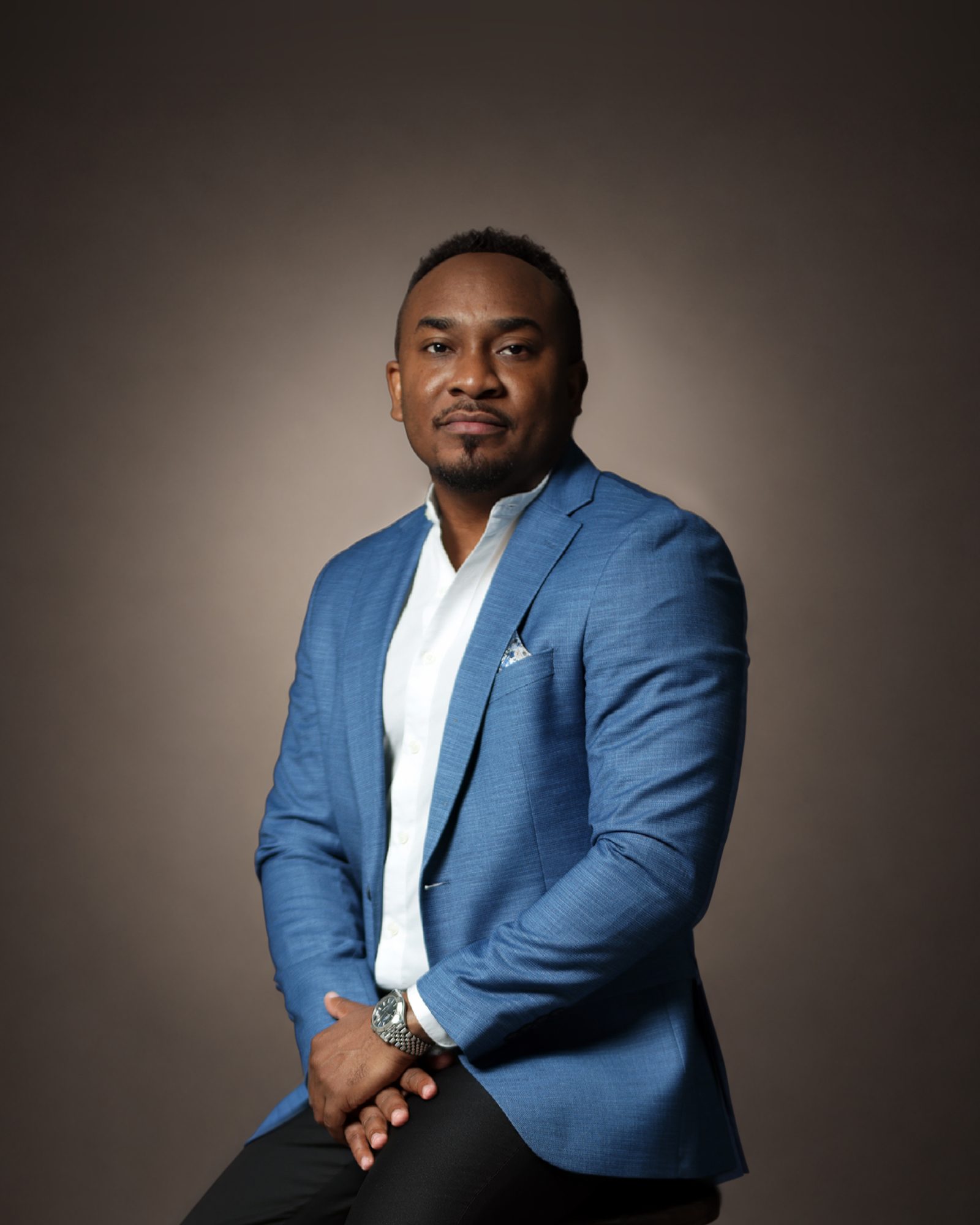 Kemal Brown in a blue blazer and white shirt, seated on a wooden stool, hands clasped, against a warm grey studio backdrop.