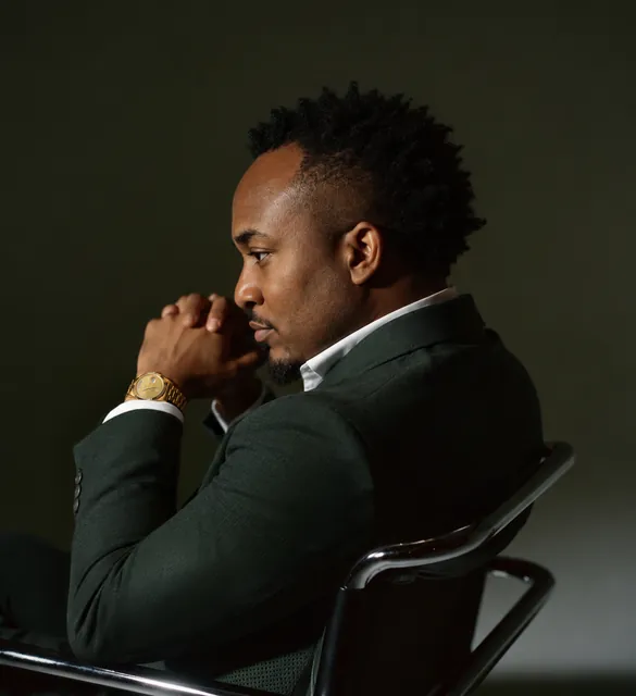 Kemal Brown in a dark green suit, seated in profile with his hands clasped at his chin.