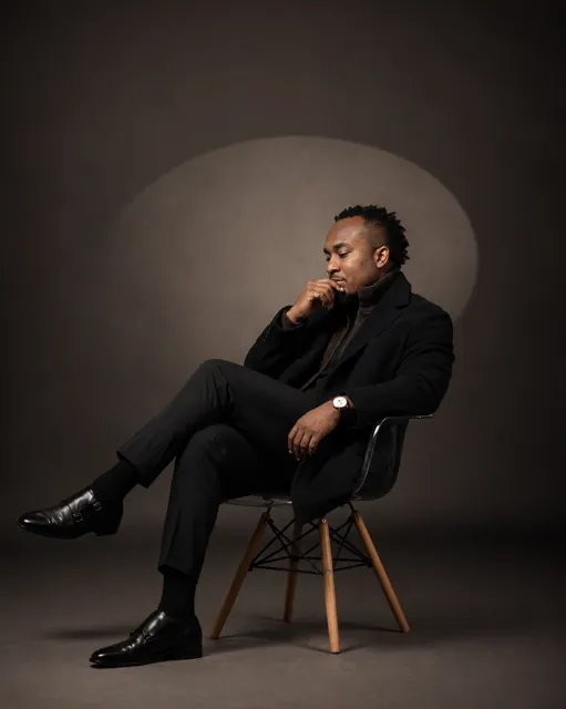 Kemal Brown in black, seated on a wooden chair in a spotlight, hand at his chin.