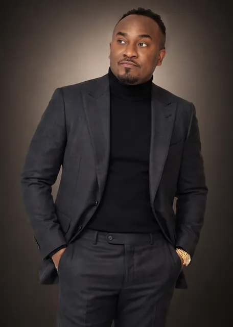 Kemal Brown in a charcoal suit and black turtleneck, hands in his pockets, in the studio.