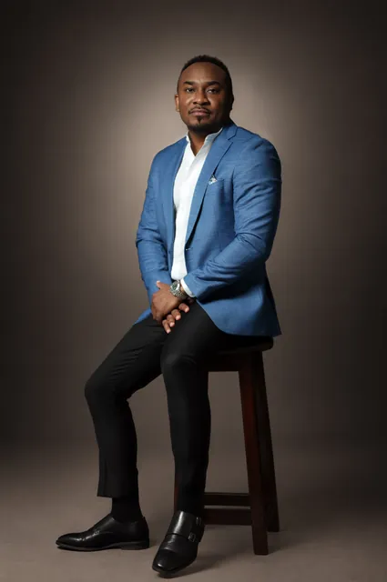 Kemal Brown in a blue blazer and white shirt, seated on a wooden stool in the studio.
